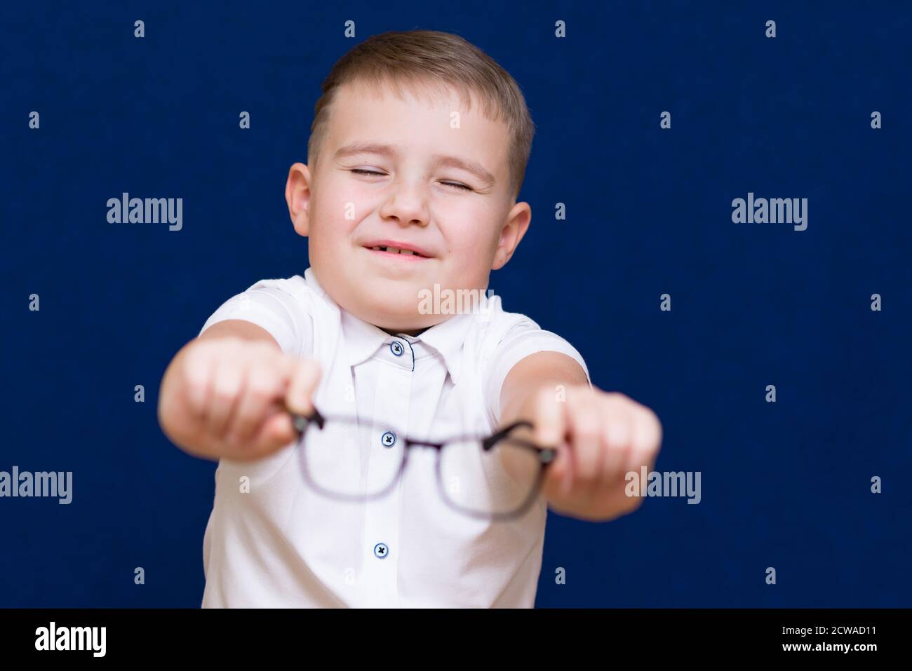 Occhi chiusi di successo 9 scolaro in piedi vicino allo sfondo blu vestito in una t-shirt bianca, tenendo e ti dà occhiali. Occhi poveri di ragazzo Foto Stock