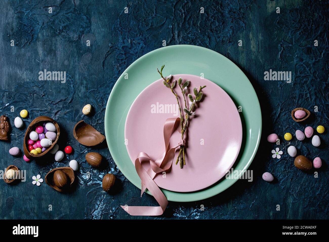 Tavola di Pasqua con uova di cioccolato, caramelle e piatti vuoti con rami di salice in fiore. Sfondo classico con texture blu. Disposizione piatta, spazio Foto Stock