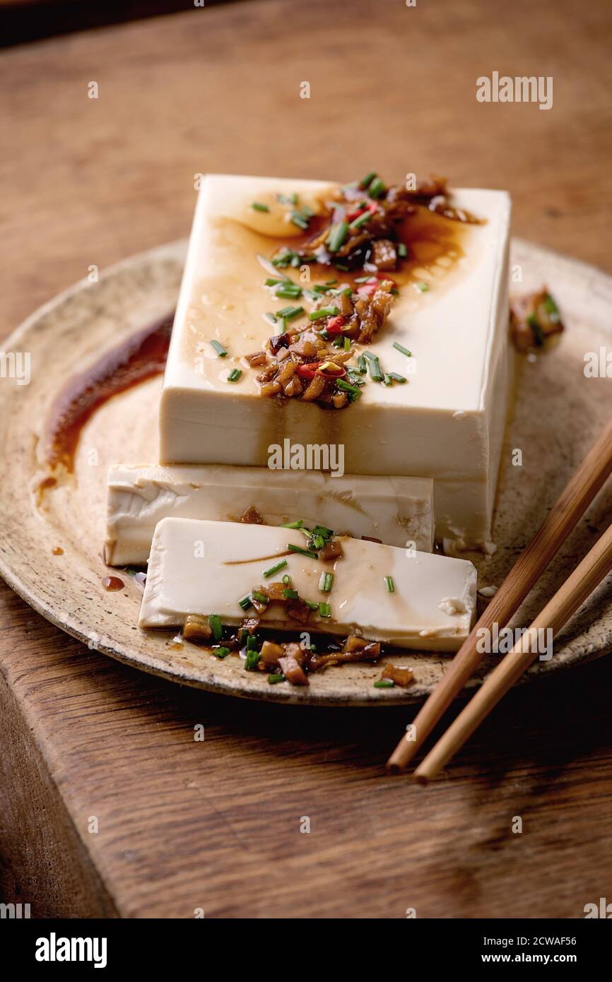 Tofu di seta giapponese formaggio di soia intero pezzo con peperoncino, zenzero in erba cipollina e salsa di soia condimento su piastra di ceramica con bastoni su tavolo di legno. Foto Stock
