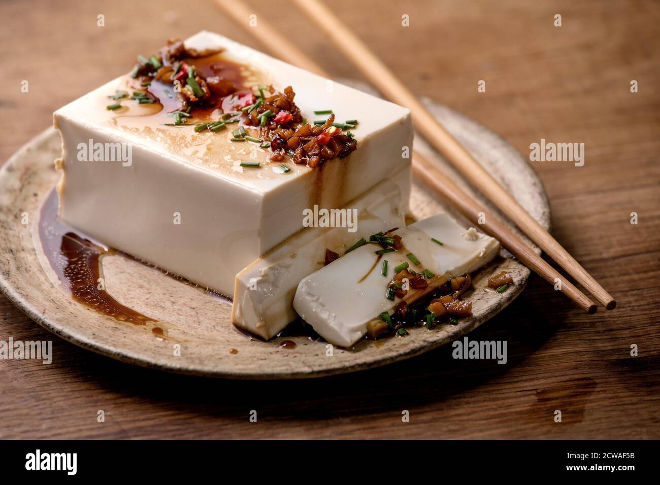Tofu di seta giapponese formaggio di soia intero pezzo con peperoncino, zenzero in erba cipollina e salsa di soia condimento su piastra di ceramica con bastoni su tavolo di legno. Foto Stock