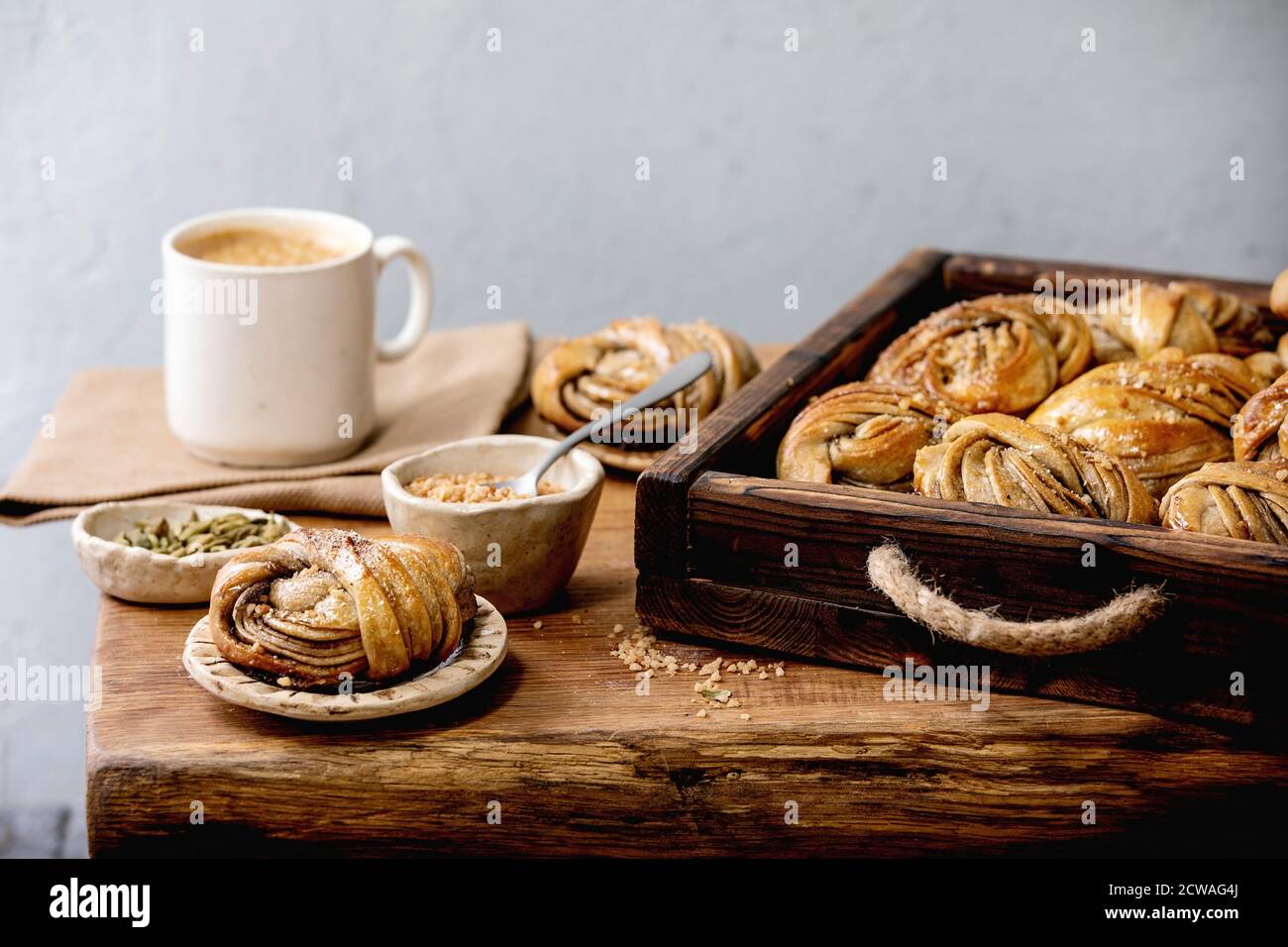 Cardamomo tradizionale svedese panini kanelbulle in vassoio di legno, tazza di caffè, ingredienti in ceramica recipiente sopra su tavolo di legno. Foto Stock