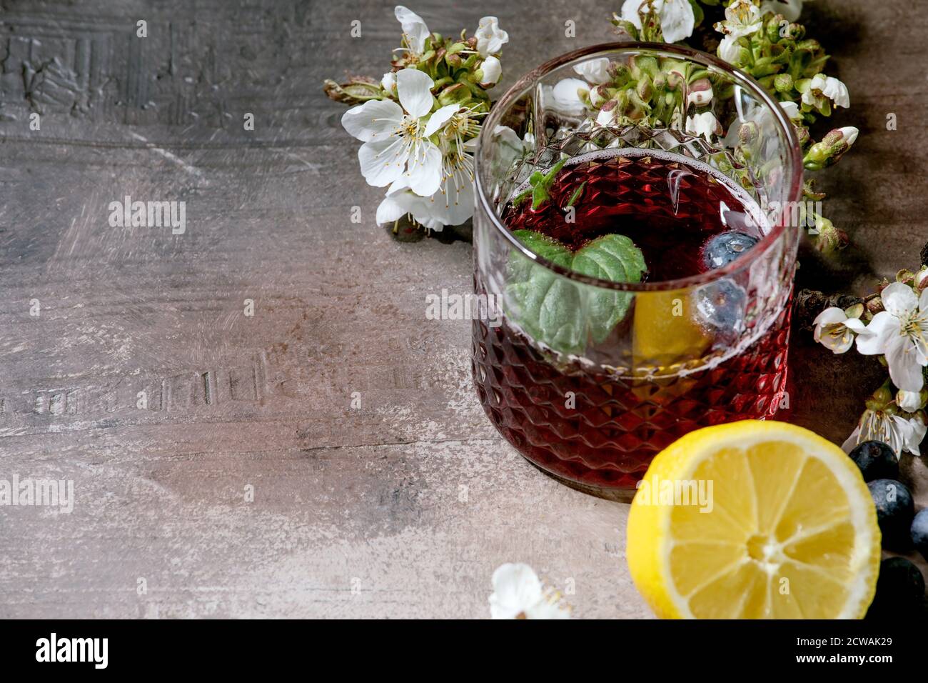 Bicchiere da cocktail con sidro frizzante al mirtillo o limonata con limone, mirtilli e menta a fette. Rami fioriti sopra. Sfondo grigio a trama. Foto Stock