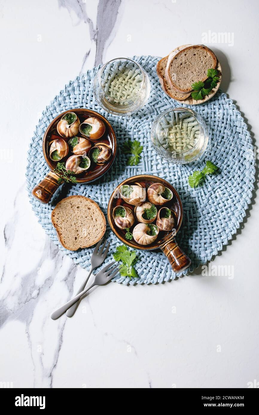 Escargots de Bourgogne - lumache con burro alle erbe, piatto gourmet, in tradizionale padella di ceramica con coriandolo, pane, bicchiere di vino bianco su tovagliolo blu Foto Stock