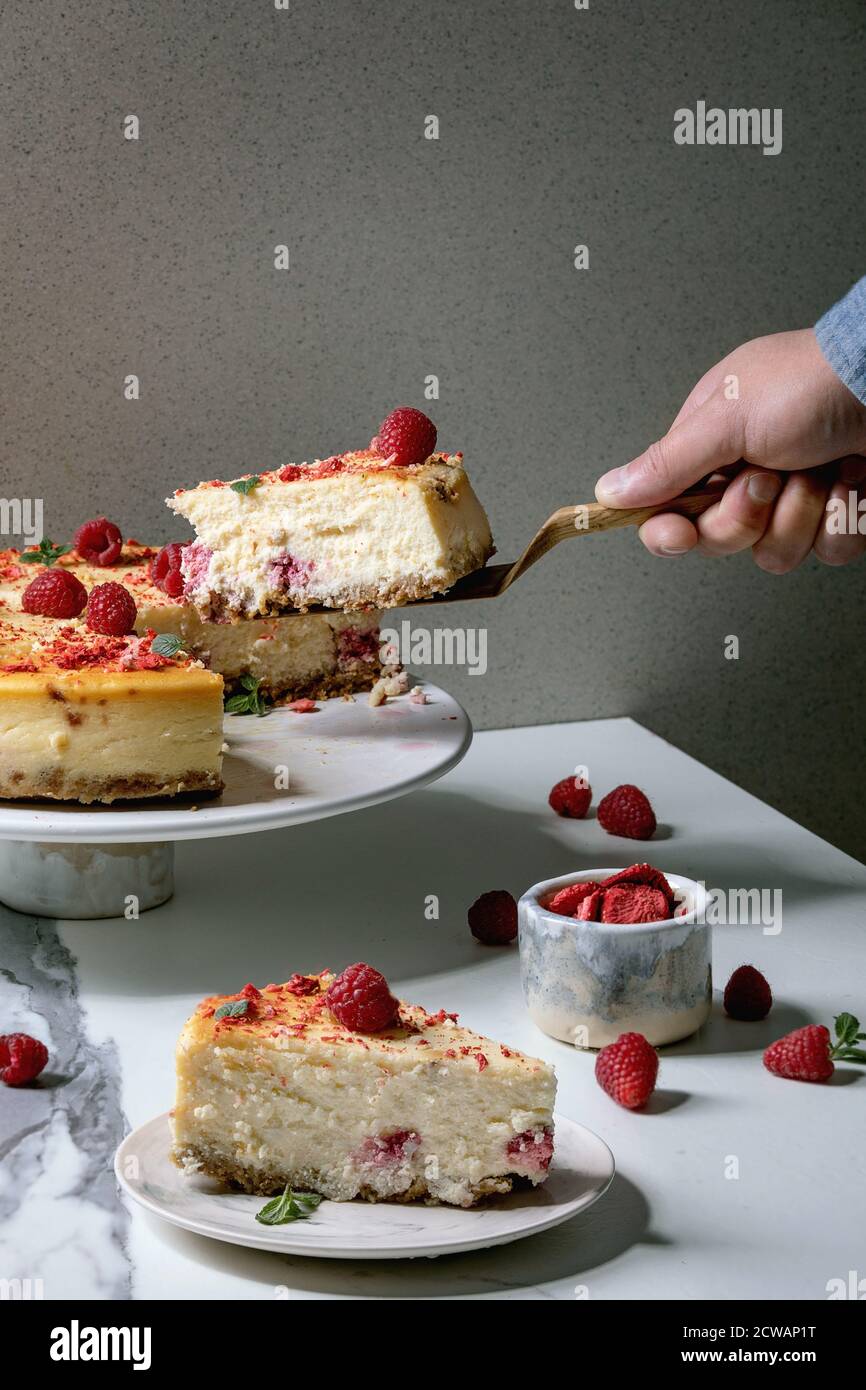 Pezzo di mano dell'uomo di lampone fatto in casa cheesecake al forno su server torta di legno. Porzione su piatto, lamponi freschi e menta sopra su marmo bianco Foto Stock