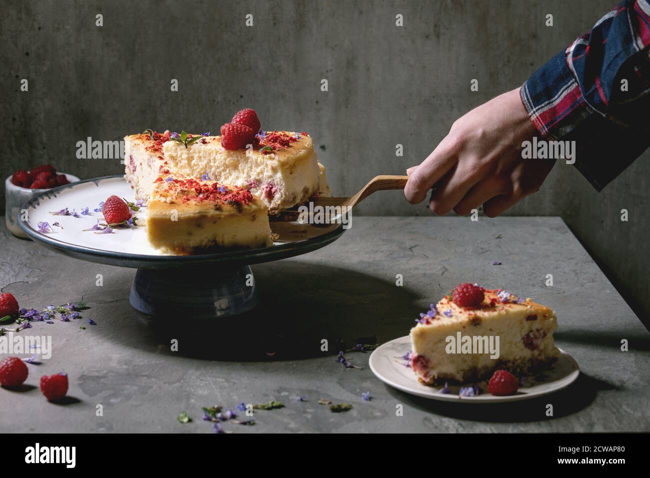 Pezzo di mano dell'uomo di lampone fatto in casa cheesecake al forno su server torta di legno. Porzione su piatto, lamponi freschi e menta sopra su tavola grigia. Foto Stock