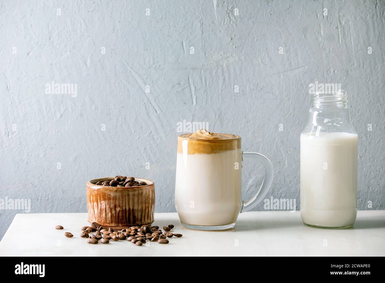 Dalgona Frothy caffè tendenza bevanda coreana latte latte latte con schiuma di caffè in tazza di vetro con ingredienti in fila su tavolo di marmo bianco. Foto Stock