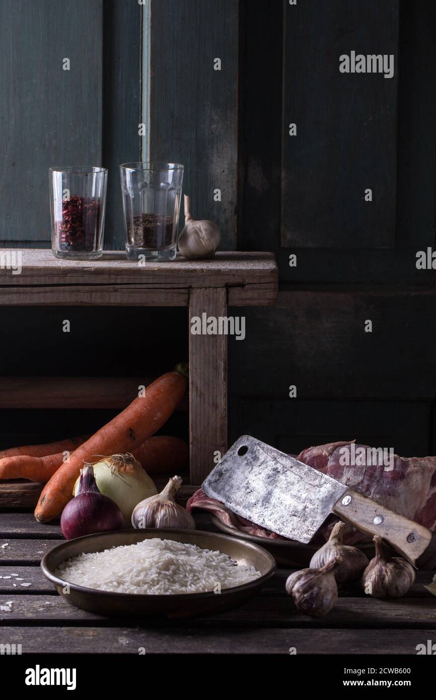 Ingredienti crudi per agnello pilaf, riso, carote, cipolla, aglio e spezie con spada di carne su tavola di legno. Stile rustico scuro. Foto Stock