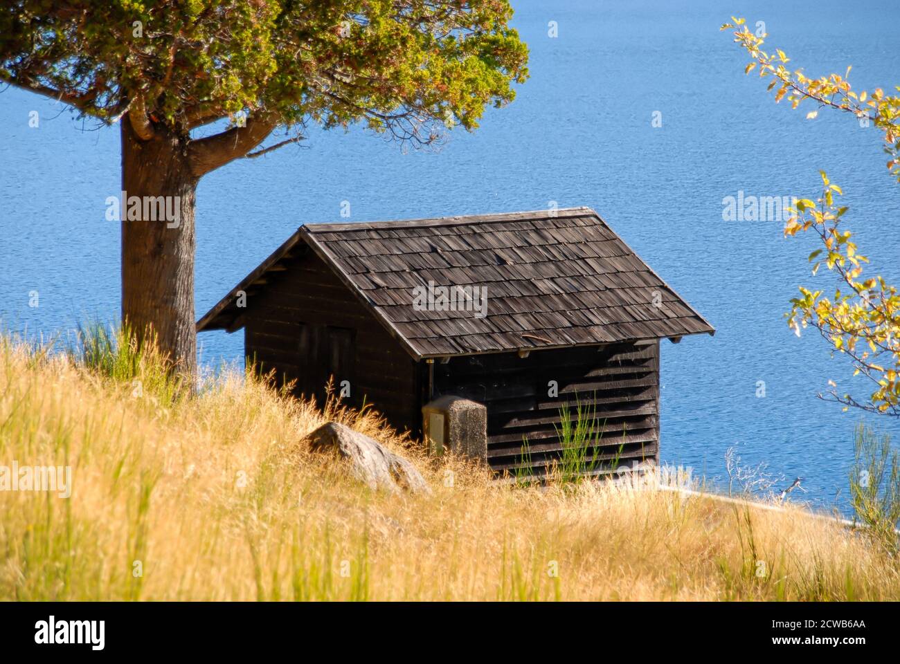 Piccolo casolare disabitato su una collina di fronte ad un lago in Argentina. Con il prato e un albero illuminato dal sole è un luogo ideale per rilassarsi Foto Stock