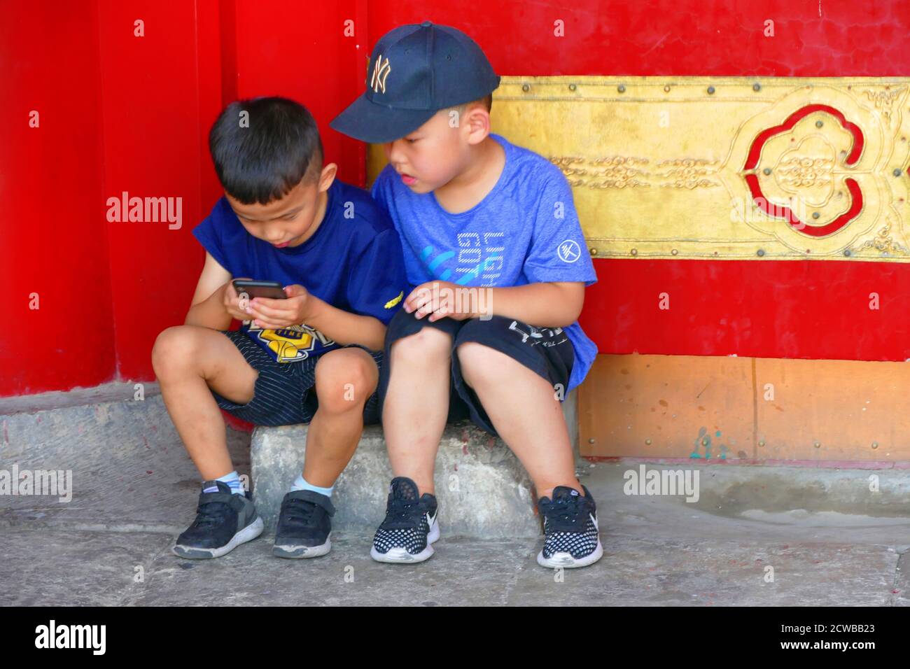 Due piccoli ragazzi giocare con un telefono cellulare al rosso cancello dorato per la Città Proibita a Pechino, Cina. La Città Proibita è un complesso di palazzo nel centro di Pechino, Cina. Essa ospita il Museo del Palazzo, e fu il primo palazzo imperiale cinese della dinastia Ming alla fine della dinastia Qing (gli anni dal 1420 al 1912). La Città Proibita è servita come casa di imperatori e le loro famiglie ed era il cerimoniale e centro politico del governo cinese per quasi 500 anni. Foto Stock