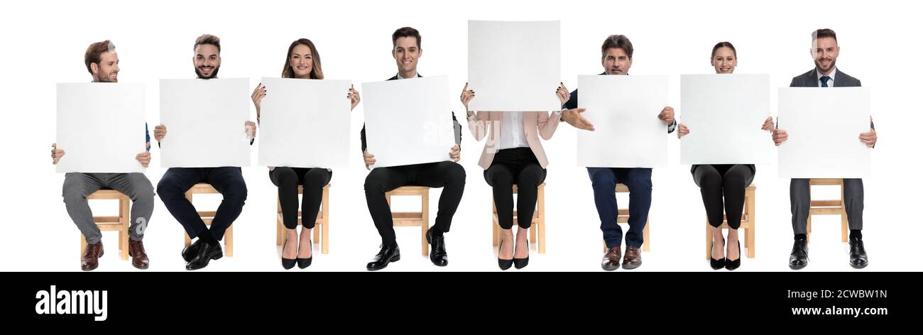 Squadra di 8 uomini d'affari che tengono i cartelloni vuoti mentre uno è nascondendo il viso mentre si siede su sedie in studio bianco sfondo Foto Stock