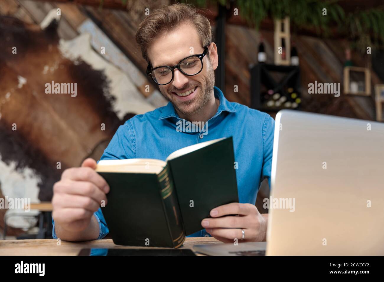 uomo informale che indossa occhiali seduti alla scrivania che studia dal libro felice al coffeeshop Foto Stock