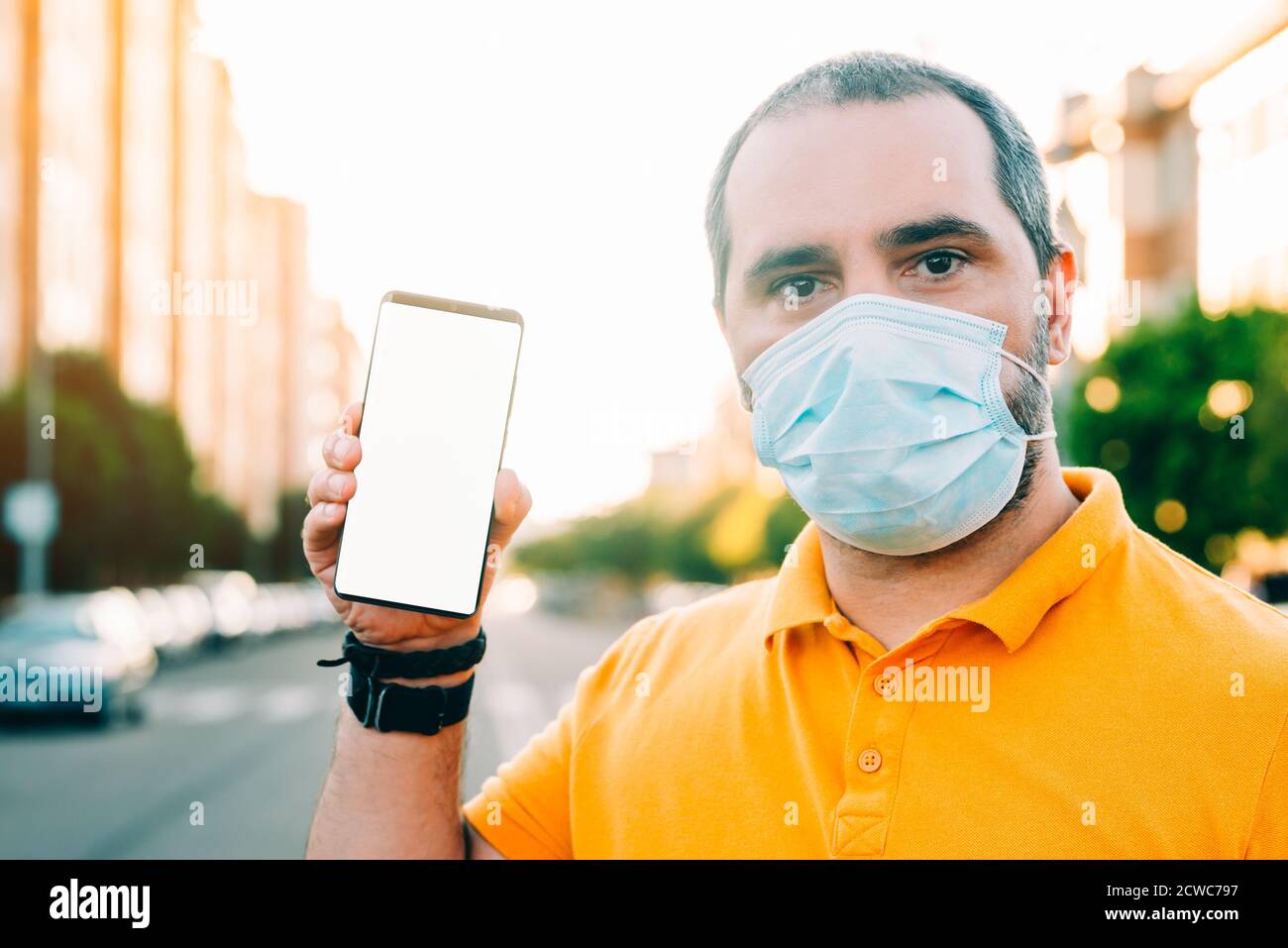 Ritratto di un uomo di 40 anni con maschera medica chirurgica in piedi, tenendo e mostrando il display dello smartphone. Foto Stock