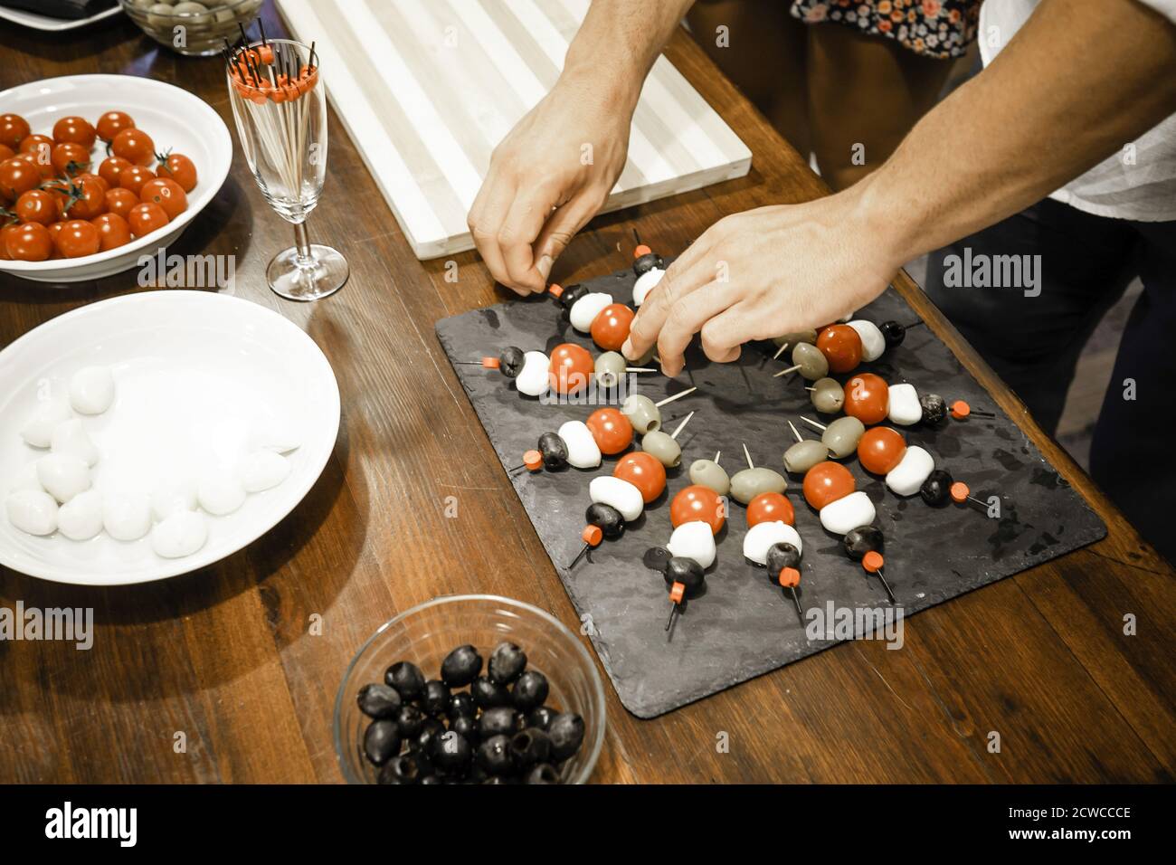 Vista dall'alto delle braccia di un giovane chef che prepara antipasti vegetariani con spiedini in un piatto d'ardesia su un tavolo di legno con mozzarella, pomodori ciliegini e o Foto Stock