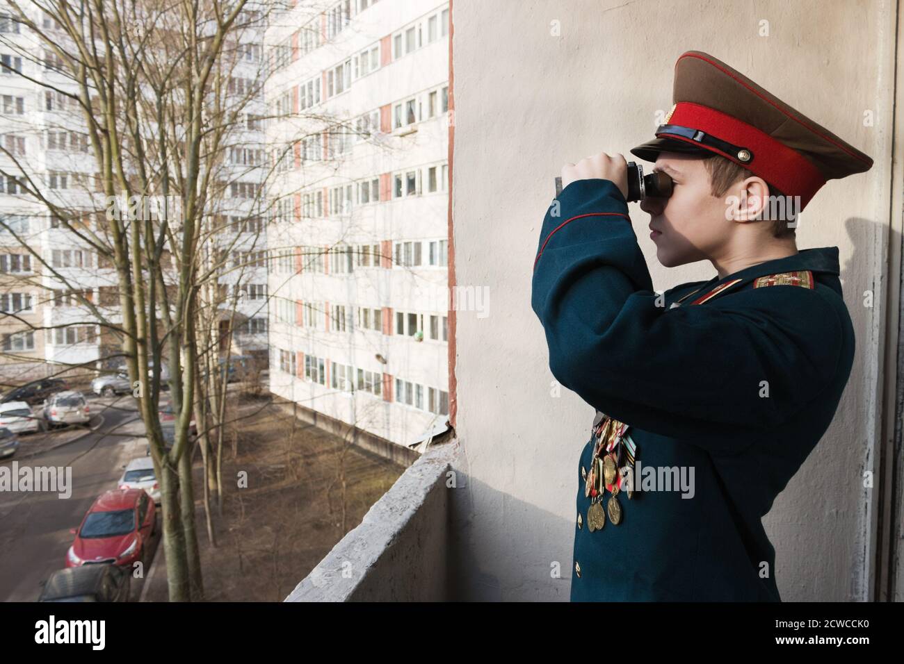Un ragazzo nella divisa completa del suo bisnonno guarda attraverso il binocolo dal balcone di un edificio residenziale Foto Stock