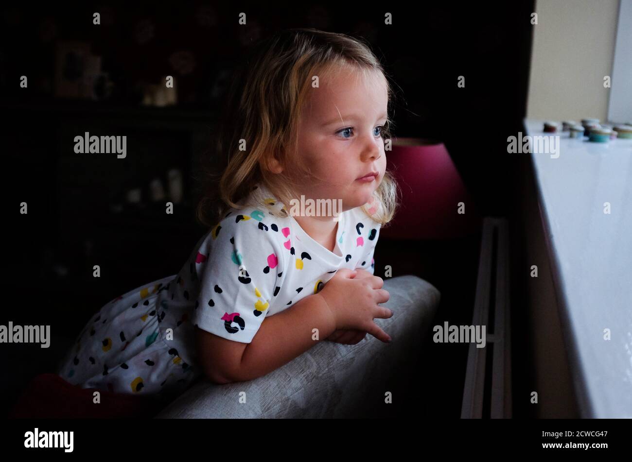 Giovane ragazza di 3 anni che guarda fuori dalla finestra con espressione pensiva - vita familiare Foto Stock