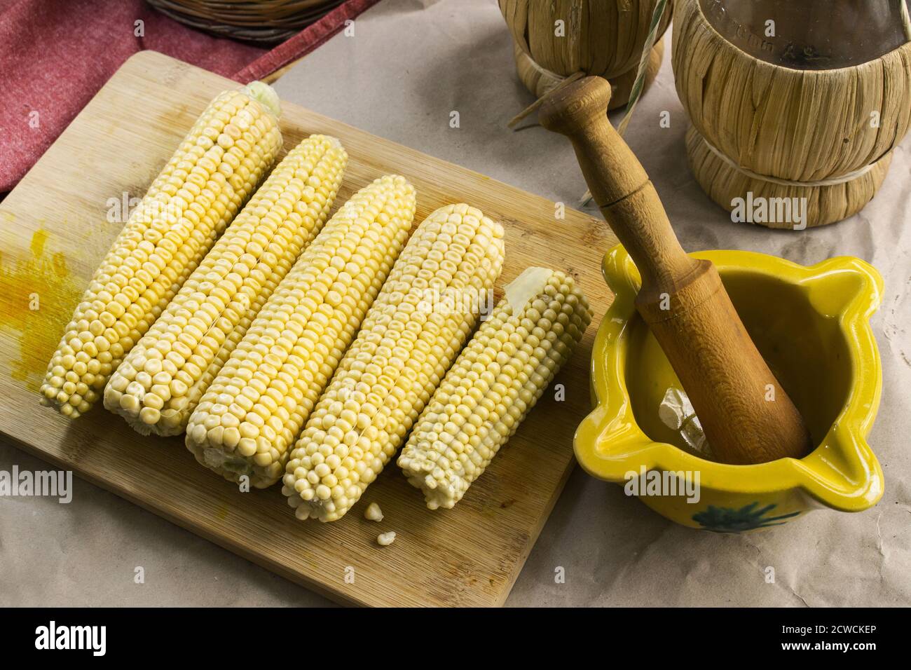 Top shot con mais crudo su un tagliere, un mortaio di ceramica valenciana e due fiasche di vimini sul fondo Foto Stock