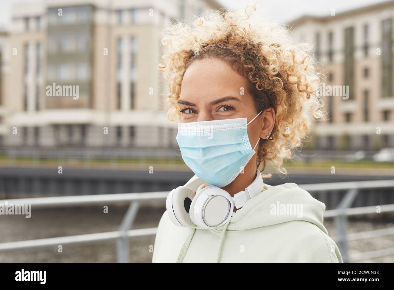 Ritratto di giovane donna con capelli ricci e maschera protettiva e guardando la macchina fotografica mentre si cammina in città Foto Stock