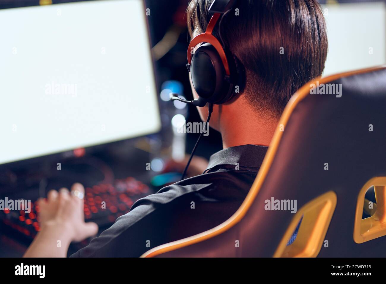 Vista posteriore di un giocatore di cybersport maschile indossando cuffie che gioca a videogiochi online mentre si siede nel club di gioco o in internet cafè. Partecipazione al torneo eSport Foto Stock