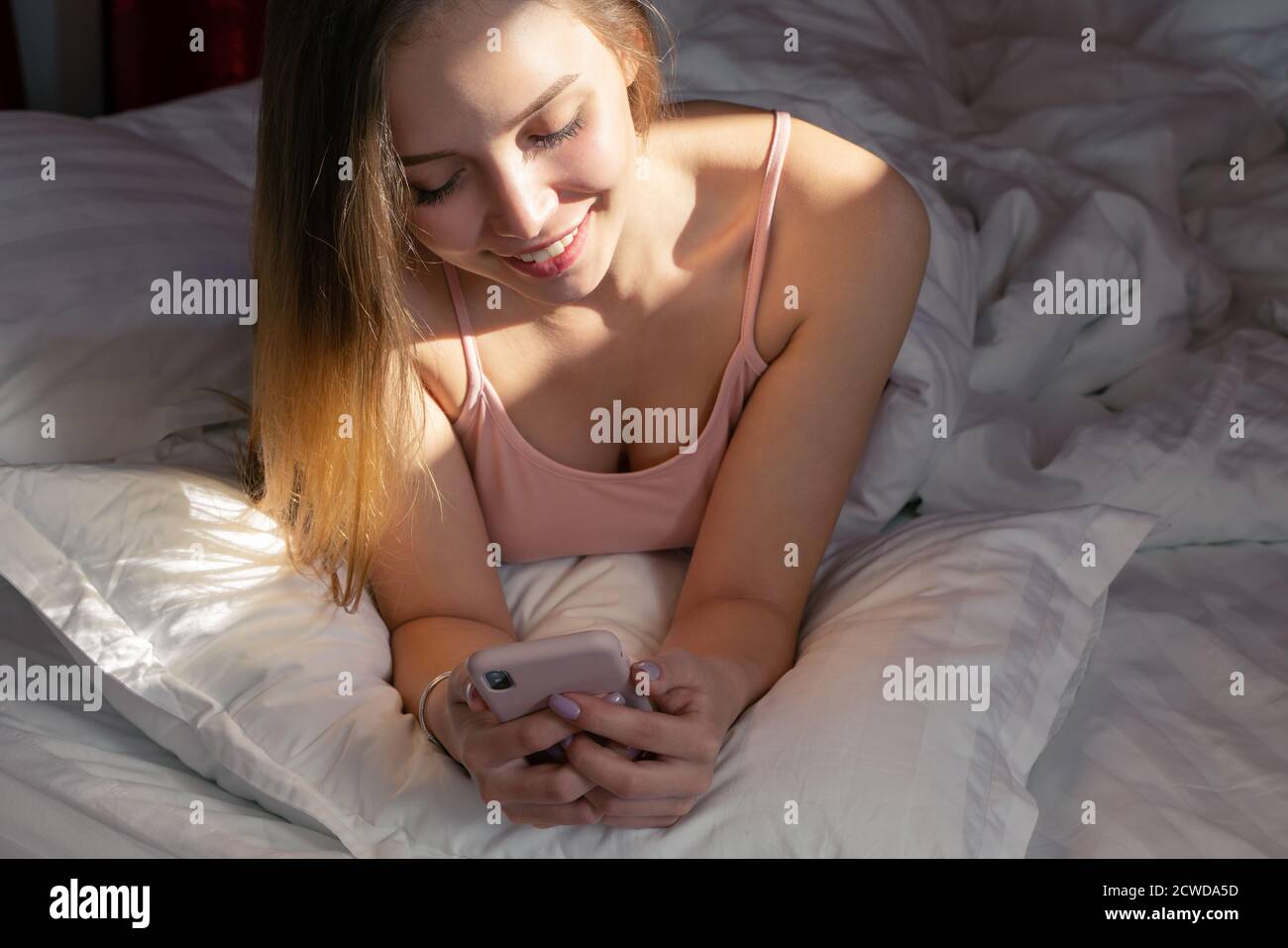 Giovane donna sorridente sdraiata nel letto bianco e che usa un telefono in camera da letto. Buongiorno. Foto Stock