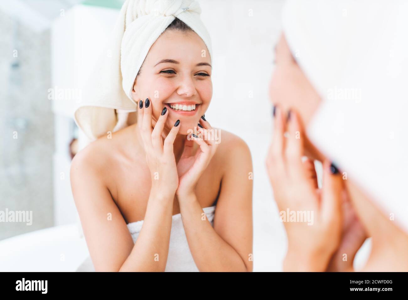 Giovane donna attraente con asciugamano bianco su testa e corpo con riflesso nello specchio in bagno. Femmina sorride e guarda se stessa Foto Stock