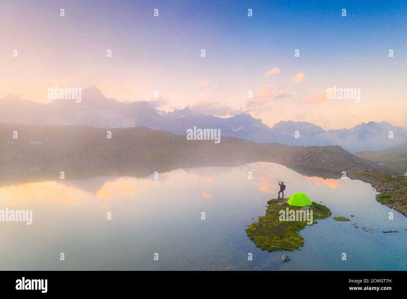 Uomo escursionista con tenda che beve da bottiglia d'acqua al lago Obere Schwarziseeli durante un alba nebbia, Passo Furka, Canton Uri, Svizzera Foto Stock