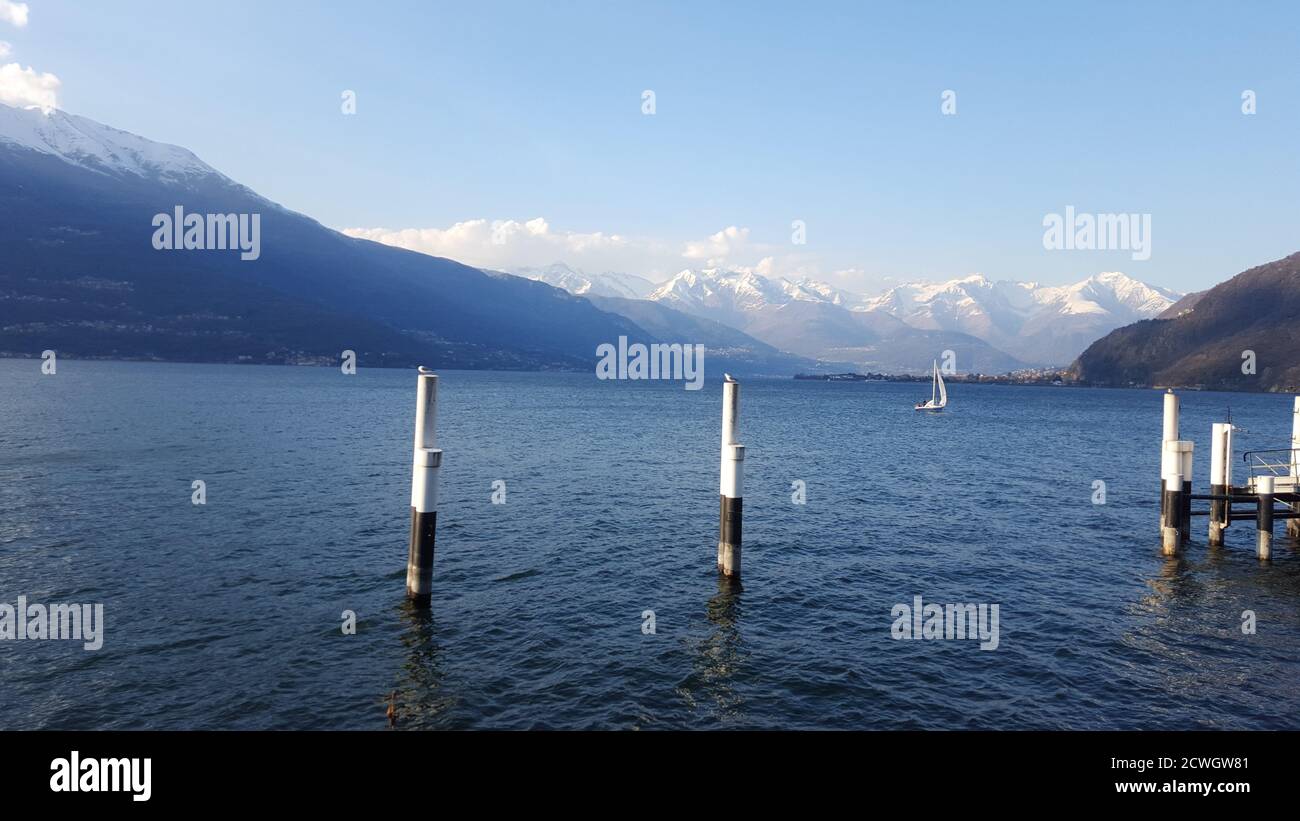 Vista panoramica invernale sui monti del Lago di Como e una barca vela in acqua dalla banchina di Bellano e due gabbiani su due pali in primo piano Foto Stock