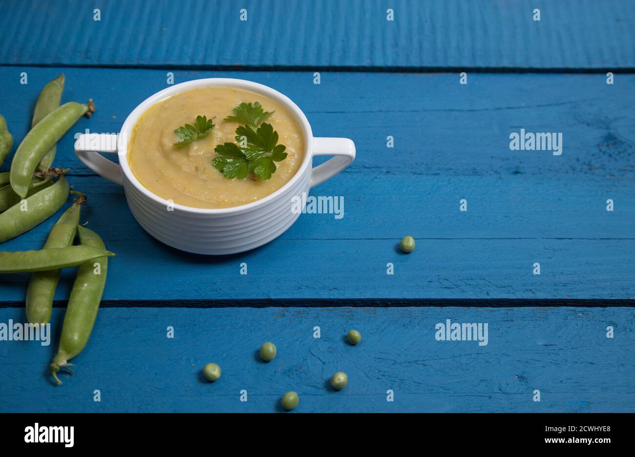 Purea di zuppa di piselli in una tazza bianca su sfondo di legno blu, accanto ai baccelli di piselli si trovano. Inserire uno spazio per la copia Foto Stock