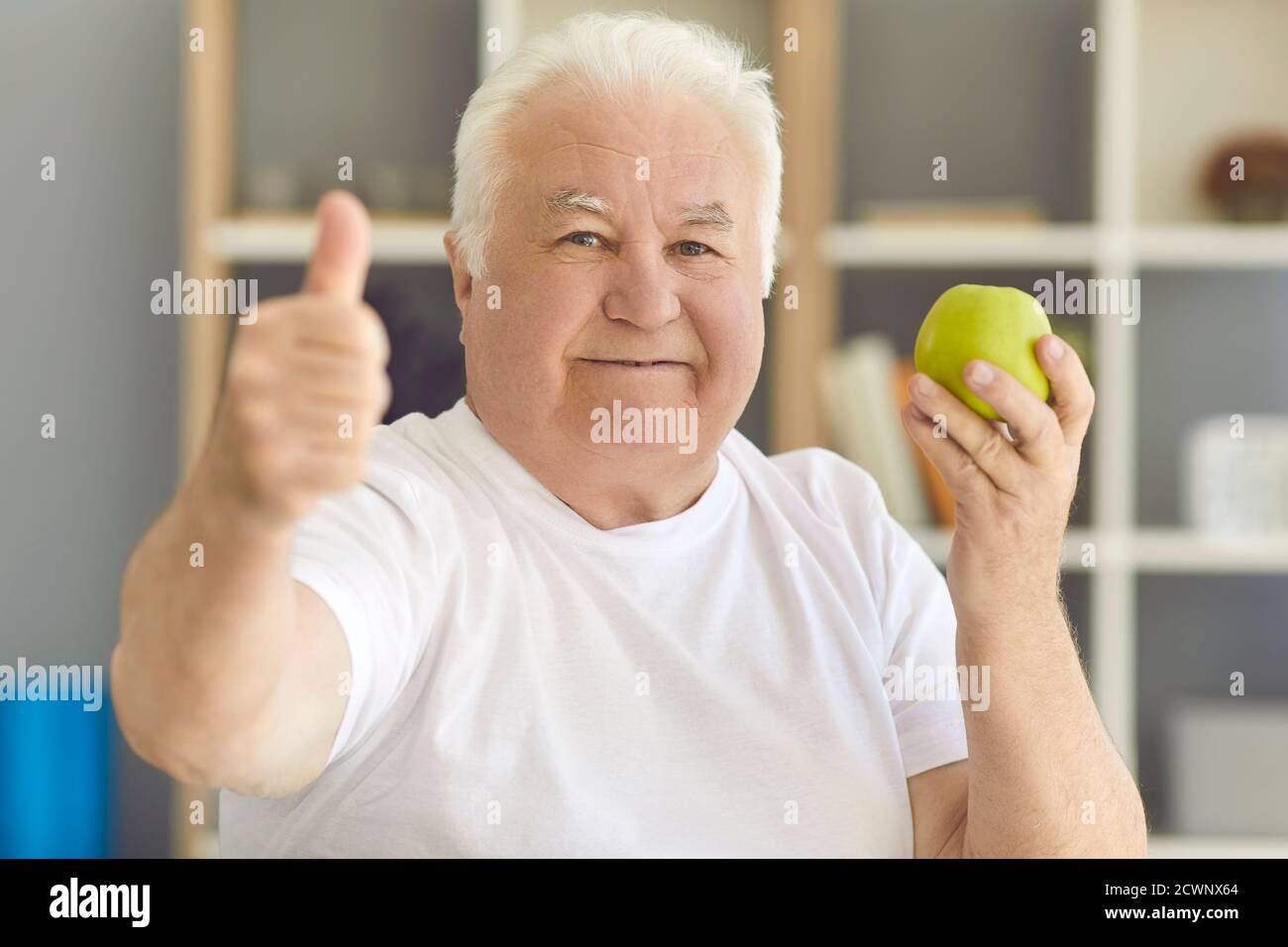 Uomo anziano felice che guarda la macchina fotografica che tiene la mela verde fresca e dando i pollici-su Foto Stock
