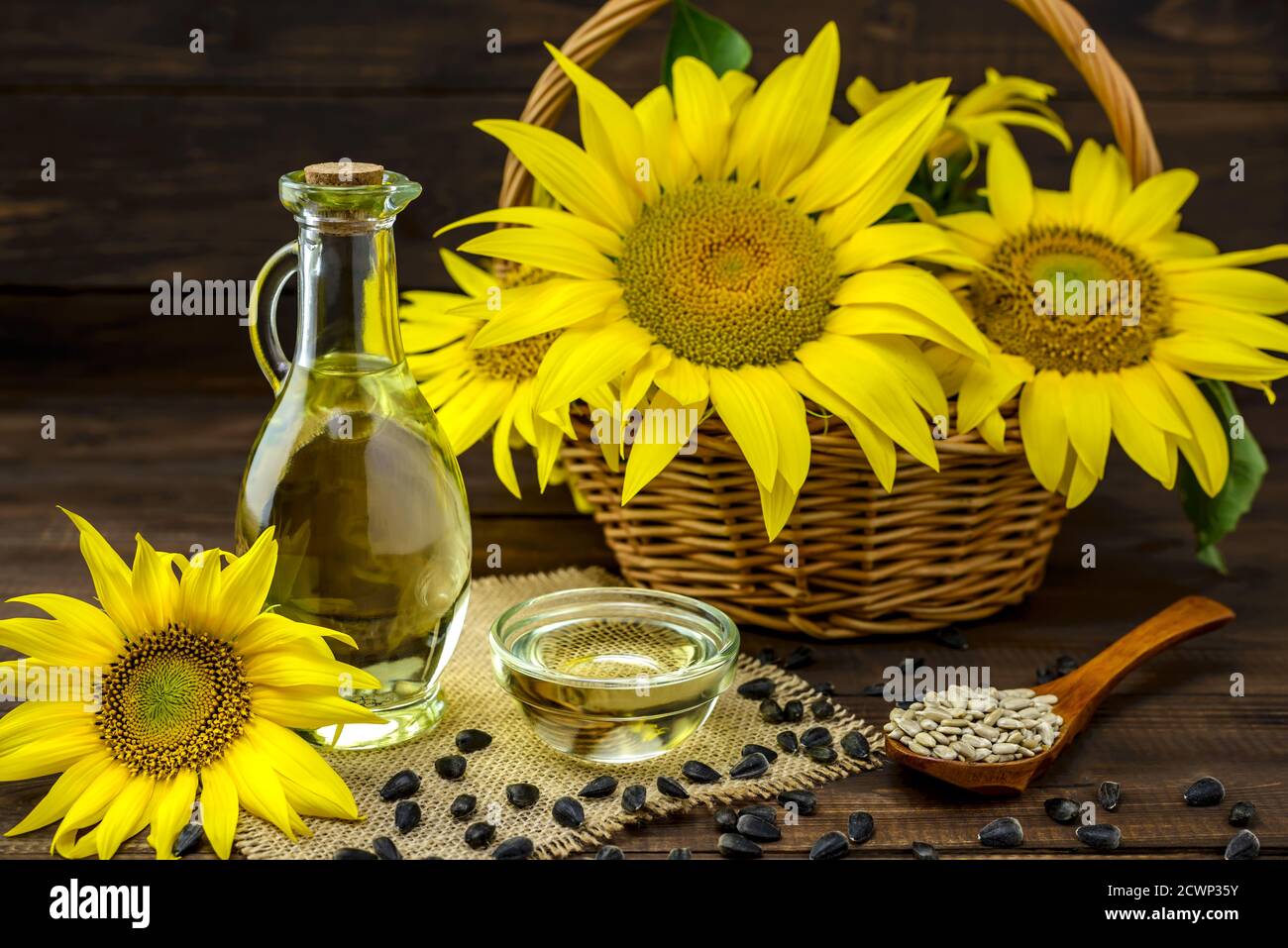 Olio di girasole in una bottiglia e semi di girasole vicino a fiori di girasole freschi in un cesto di vimini su sfondo di legno. Messa a fuoco selettiva. Foto Stock