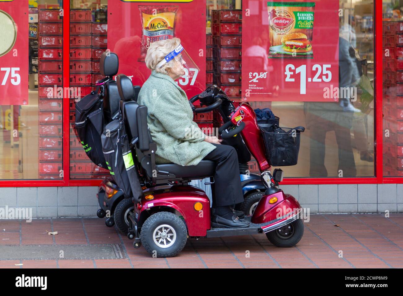 Una signora anziana che indossa uno scudo sul viso seduto su un scooter di mobilità fuori da un supermercato durante la pandemia del coronavirus Foto Stock