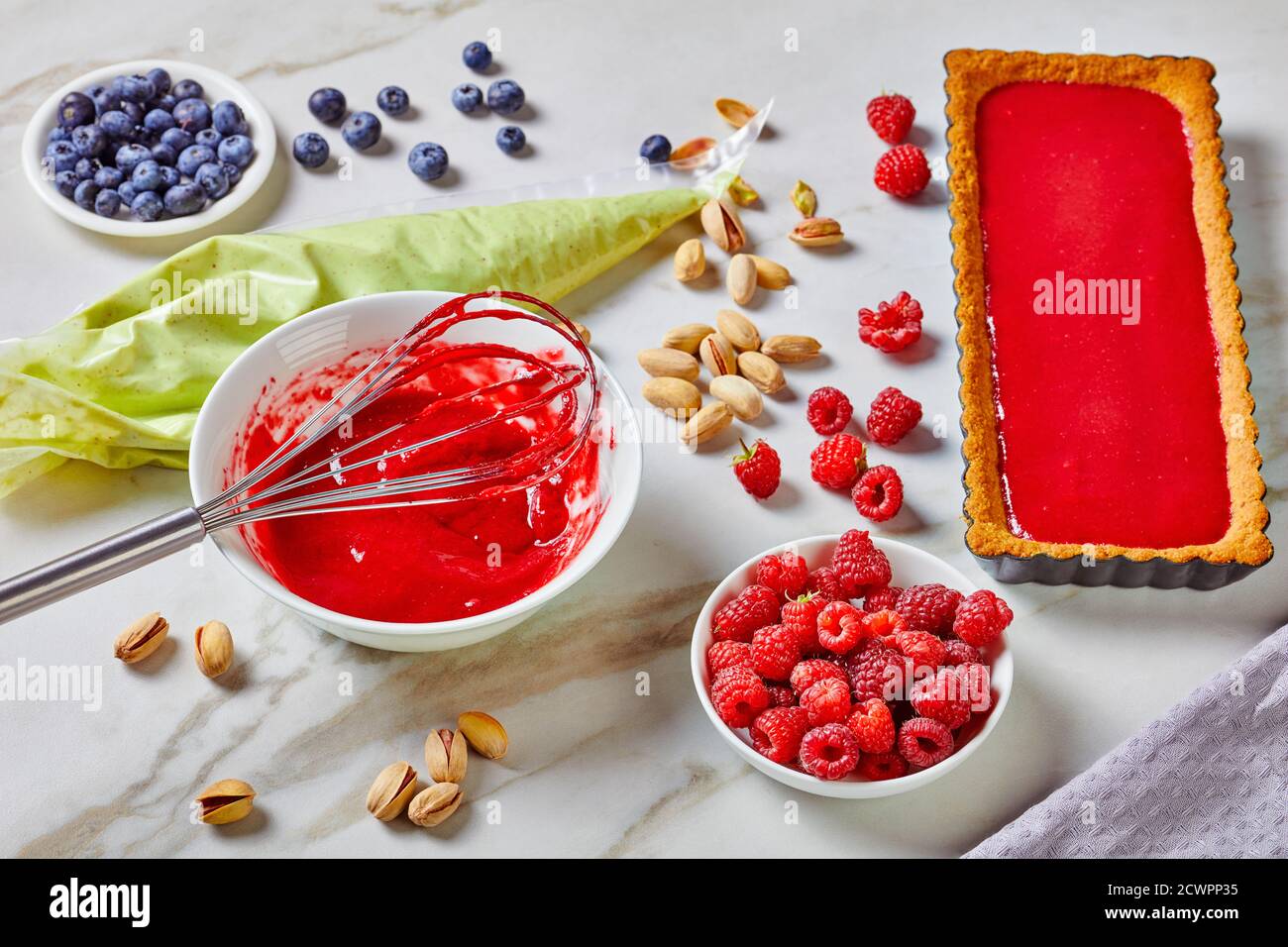 Crostata di pistacchio ganache con gelatina di pectina di lampone servita su sfondo marmoreo con frusta, sacchetto di pasticceria ripieno di cioccolato bianco e pistacchio ga Foto Stock