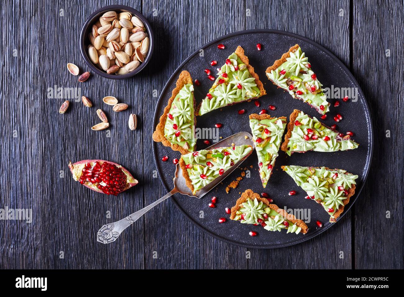 Crostata di ganache al pistacchio con gelatina di melograno, semi di melograno sulla parte superiore, servita su un piatto nero servito su un tavolo di legno rustico scuro con panino da dessert Foto Stock