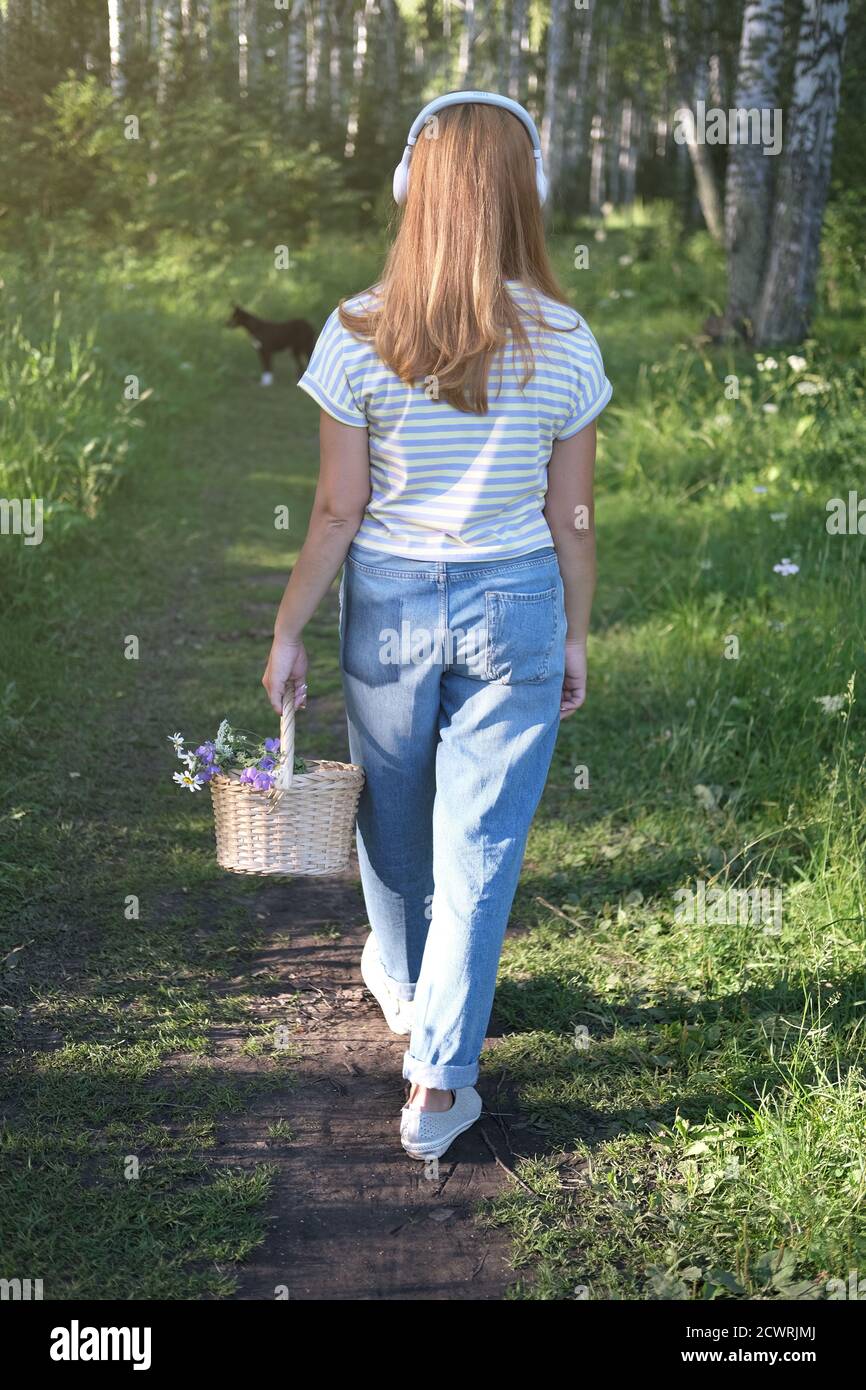 bella donna di mezza età che cammina nella foresta con cesto di fiori e un cane. passeggiata serale estiva. vista posteriore di una ragazza con cuffie. dimensioni verticali. Foto Stock