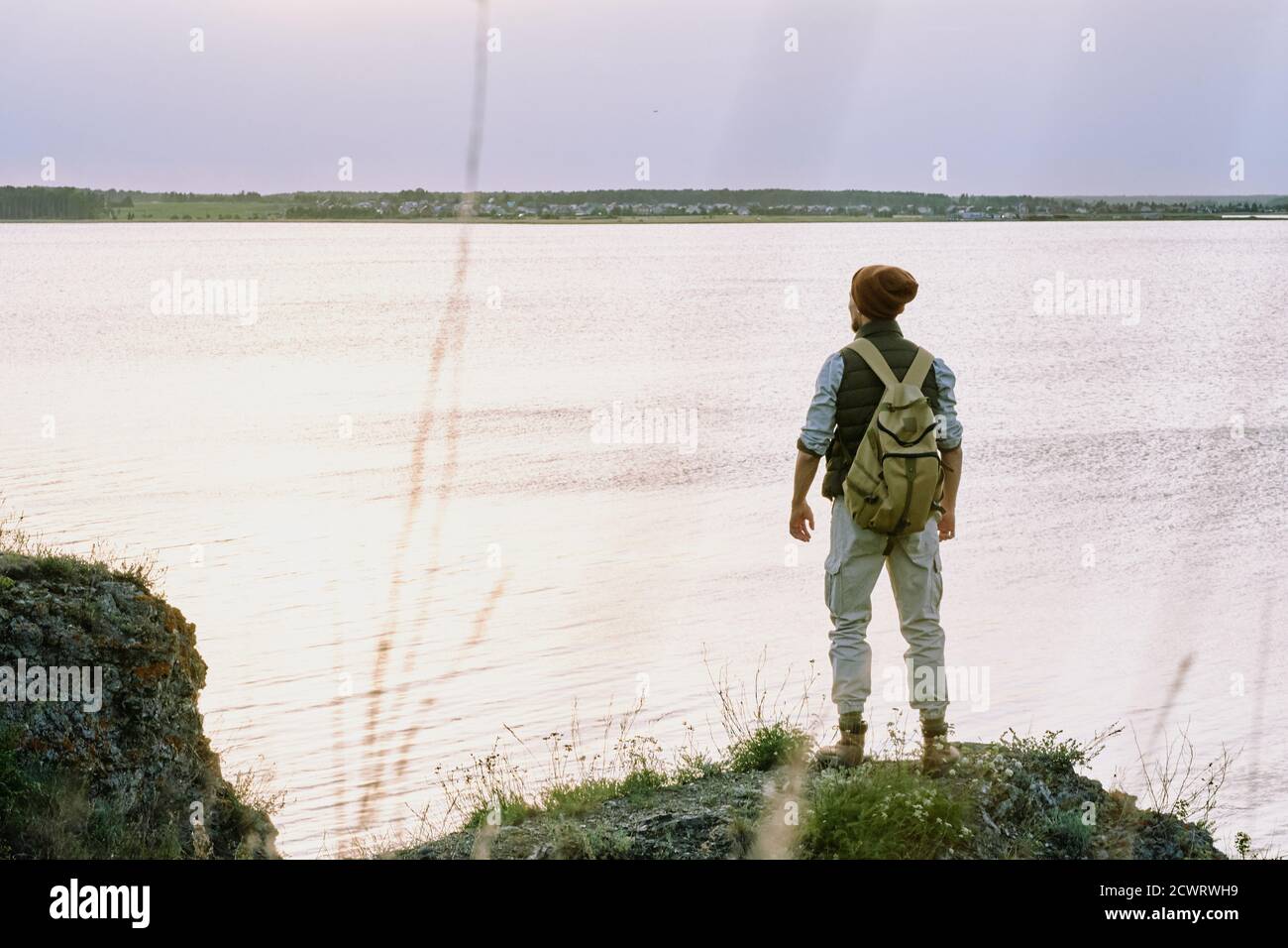 Vista posteriore di un giovane zaino in spalla maschio in cappello in piedi bordo della scogliera e contemplare il lago mentre ti godi un'escursione Foto Stock