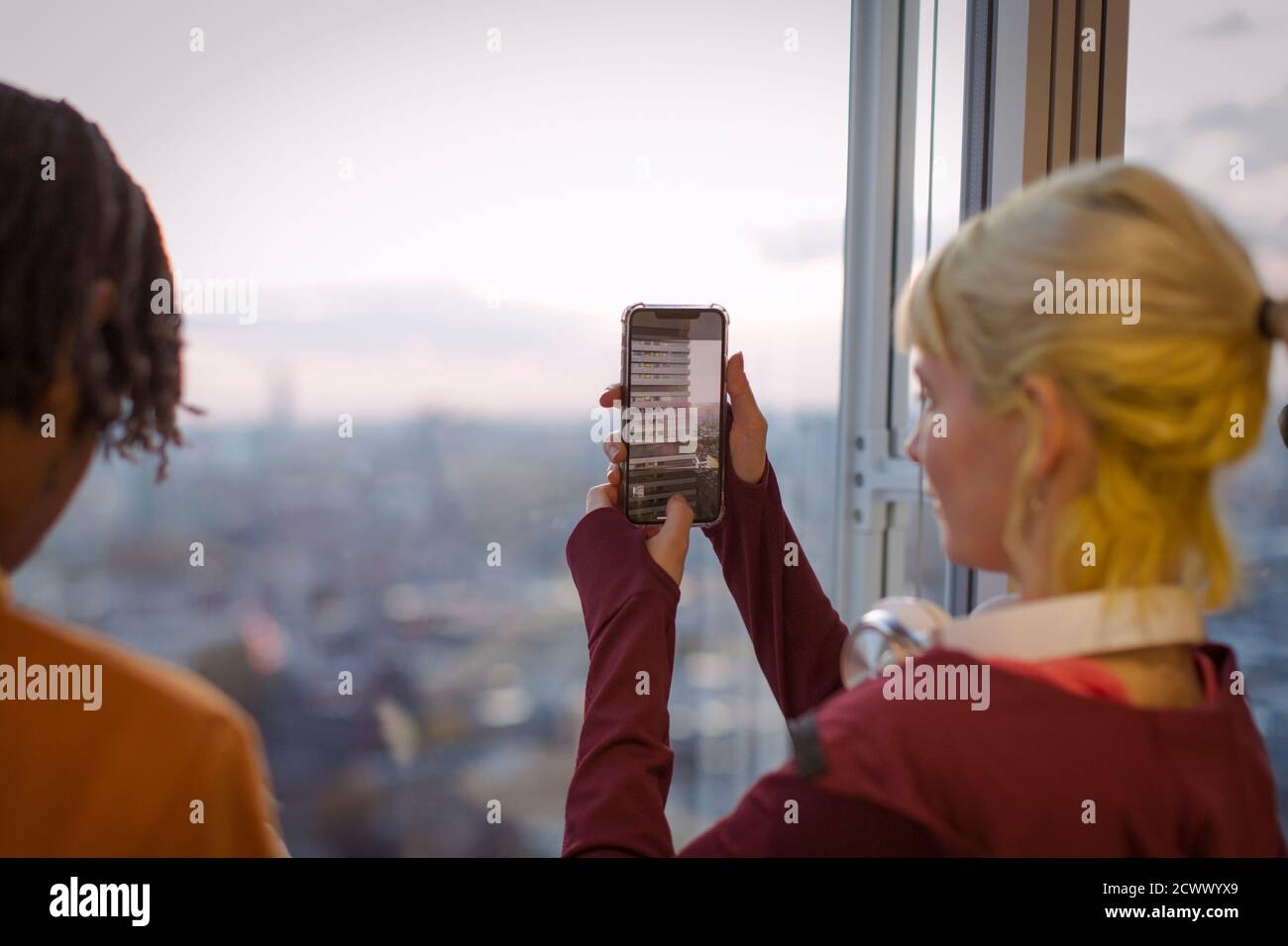 Giovane donna che usa il telefono della macchina fotografica alla finestra dell'ufficio di Highrise Foto Stock