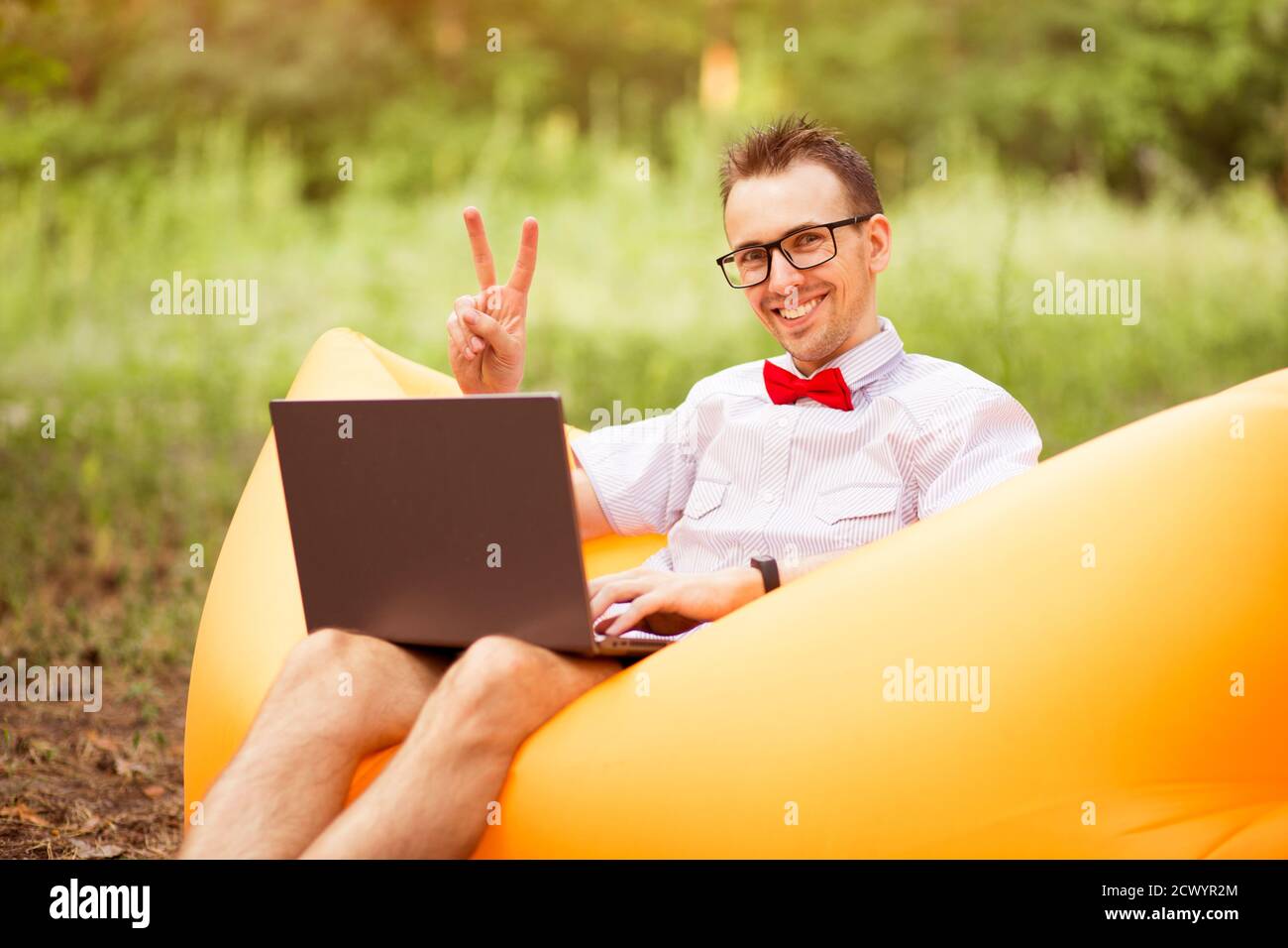 Ritratto di bel successo positivo bearded giovane freelancer in camicia, cravatta arco rosso e occhiali mostra v gesto e seduta su divano gonfiabile con Foto Stock