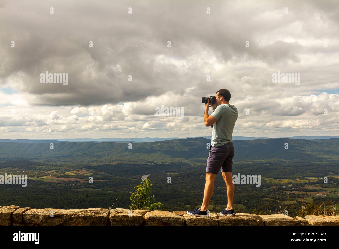 Un giovane fotografo di paesaggio caucasico che indossa una camicia corta e una t-shirt Sta scattando una foto della valle di Shenandoah sulla parte superiore di una parete di barriera di pietra da una scogliera Foto Stock