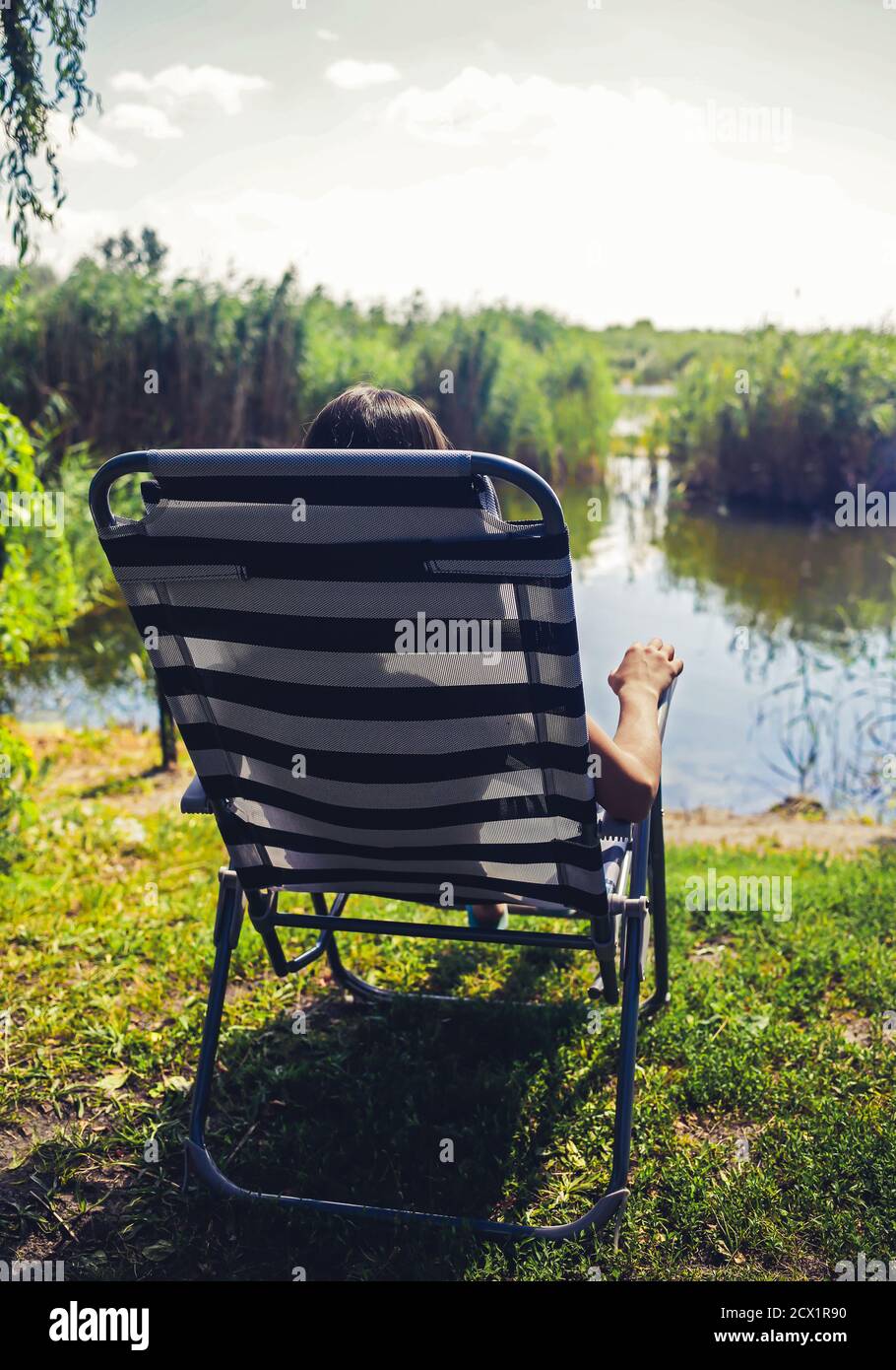 La ragazza si siede e si rilassa al lettino a strisce, guardando il piccolo lago con uno sfondo verde natura bella Foto Stock
