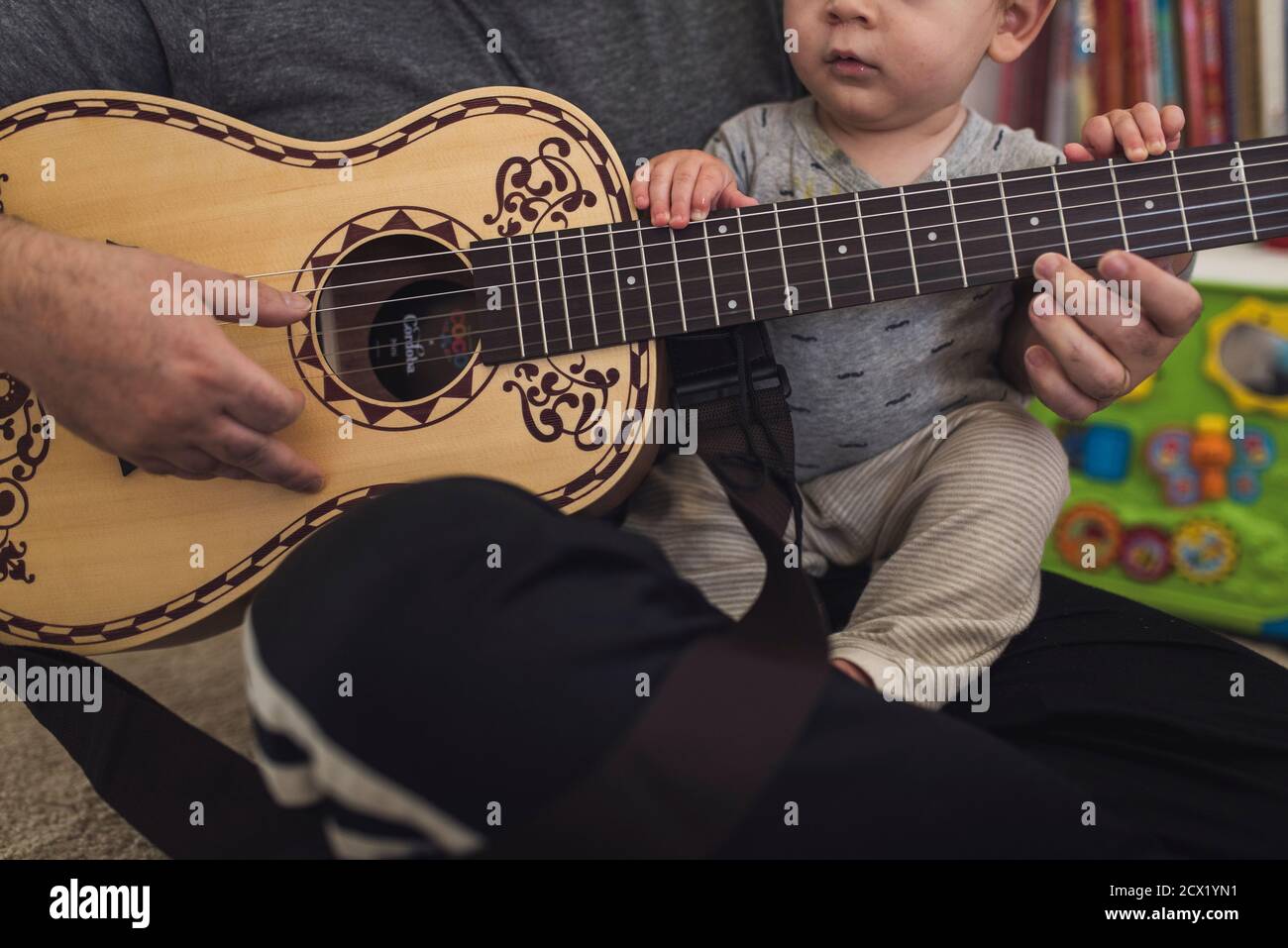 Papà sul pavimento che suona una chitarra di dimensioni ridotte tenendo in mano 1 anno di età. Foto Stock