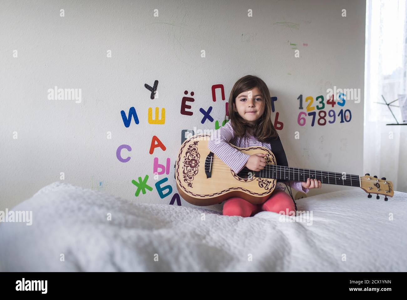 bambina di 6 anni con chitarra sul letto davanti Di muro con alfabeto greco Foto Stock