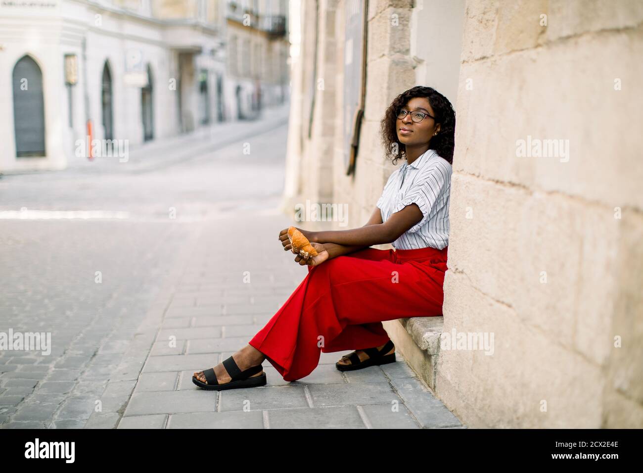 Foto orizzontale di giovane e attraente donna africana sognante vestita di pantaloni  rossi e camicia leggera, seduto sulle scale del vecchio edificio d'epoca  sul Foto stock - Alamy