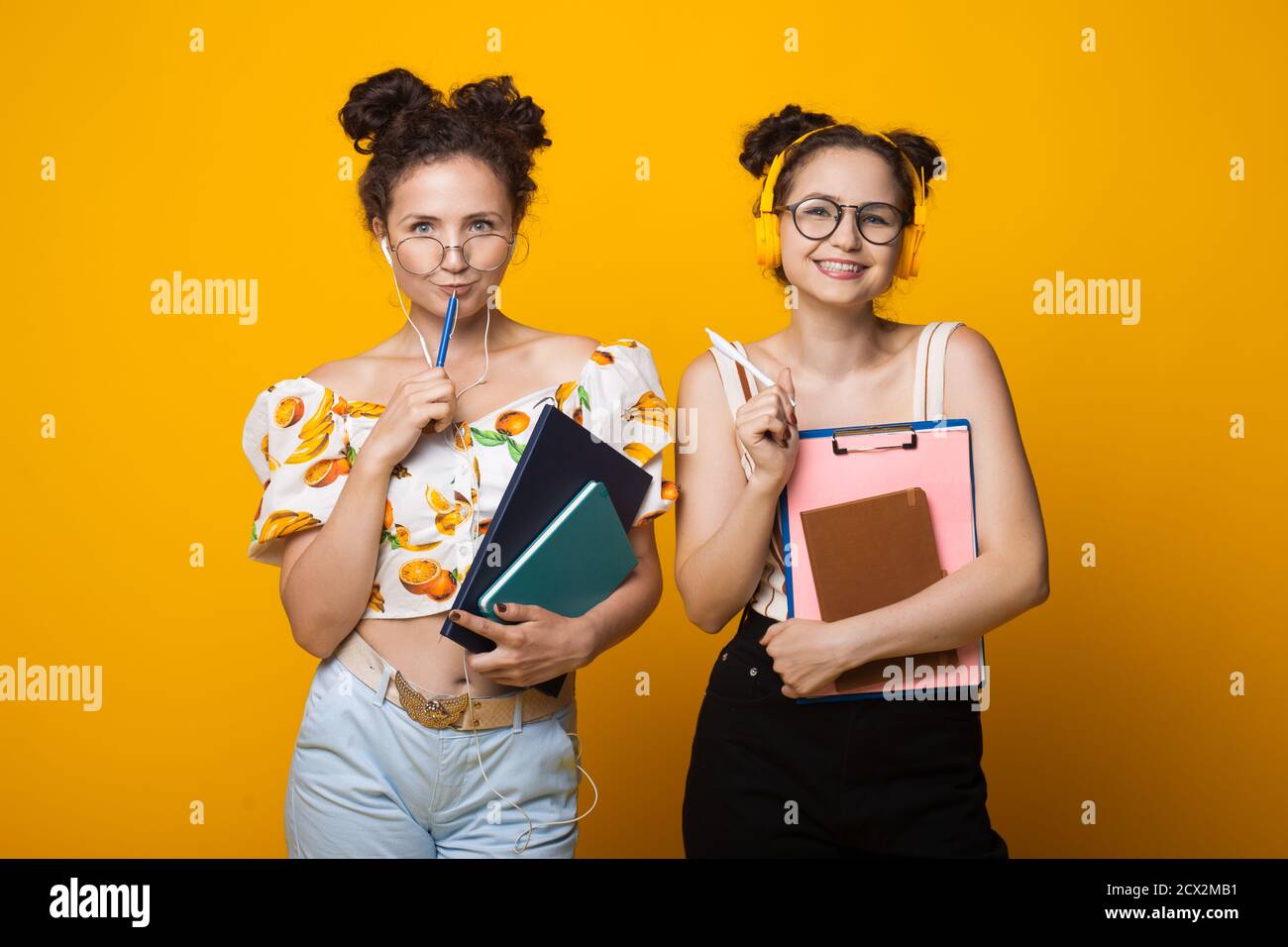 I gemelli dai capelli ricci sorridono alla macchina fotografica attraverso gli occhiali che indossano gli auricolari e tenendo alcune cartelle su una parete gialla Foto Stock