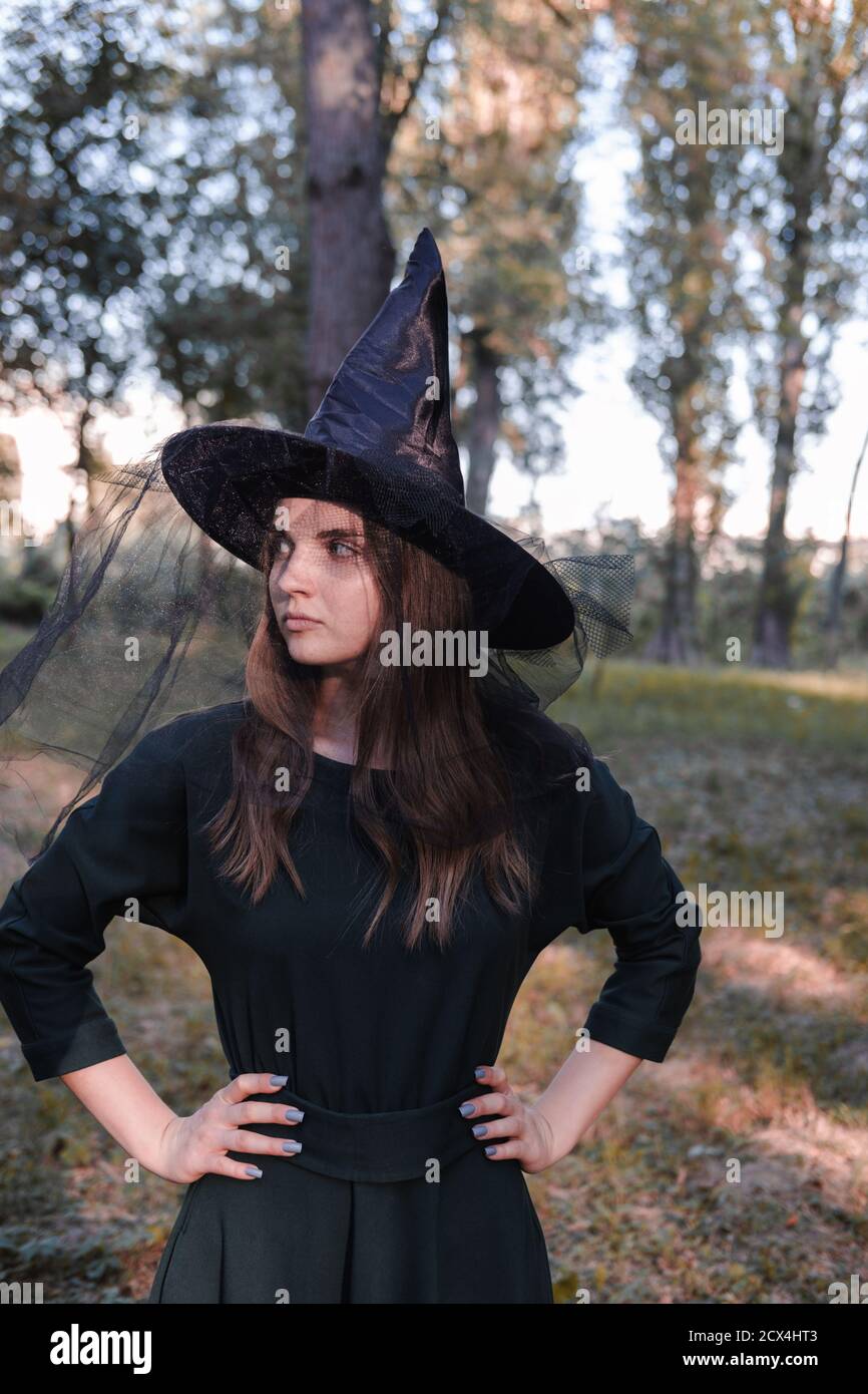 Bella donna giovane bella bella in abito scuro e cappello di strega in piedi nel mezzo dei boschi o parco autunnali. Costume da festa di Halloween. Primo piano portrai Foto Stock