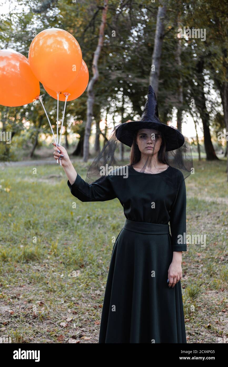 Giovane bella donna in abito scuro e cappello di strega tiene palloncini arancioni nelle sue mani. Costume da festa di Halloween. Foresta, parco con alberi d'autunno Foto Stock