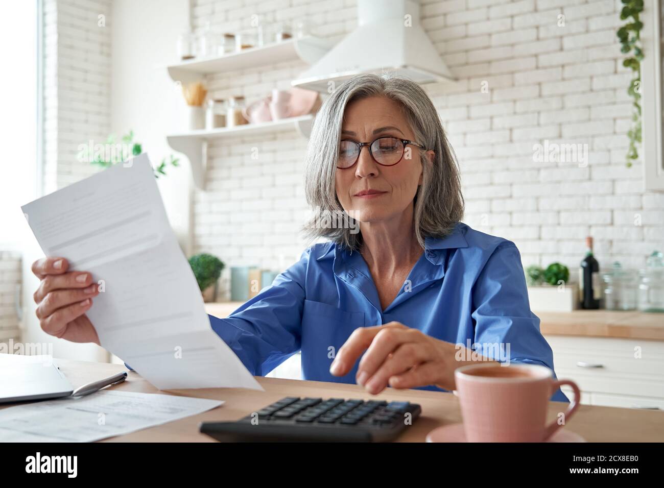Donna matura che tiene la fattura della carta usando la calcolatrice che gestisce le finanze a casa. Foto Stock