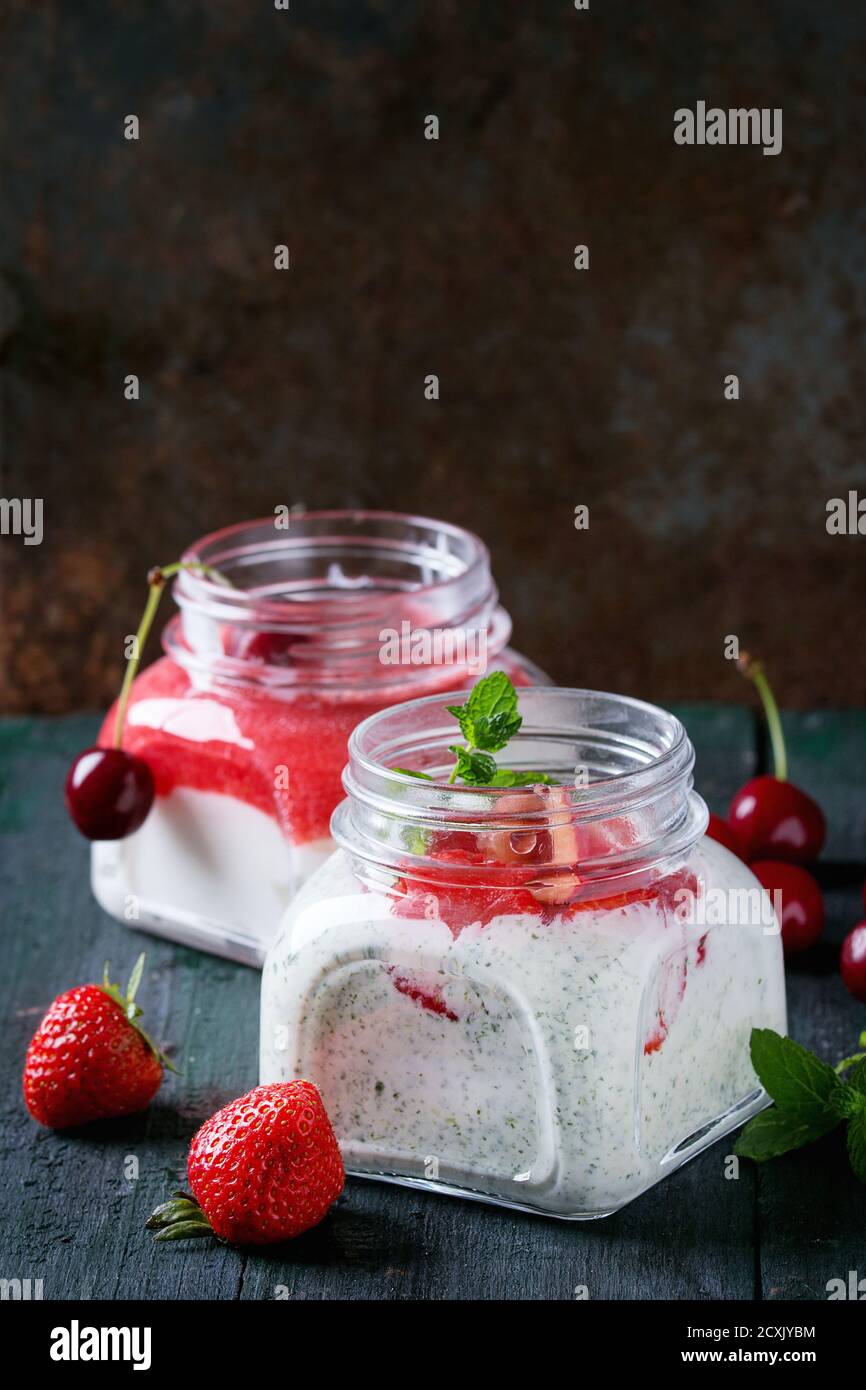 Due vasetti di vetro quadrati con yogurt fatto in casa con menta, purea di fragole e ciliegia, serviti con frutti di bosco freschi su sfondo di legno scuro. Con spazio f Foto Stock