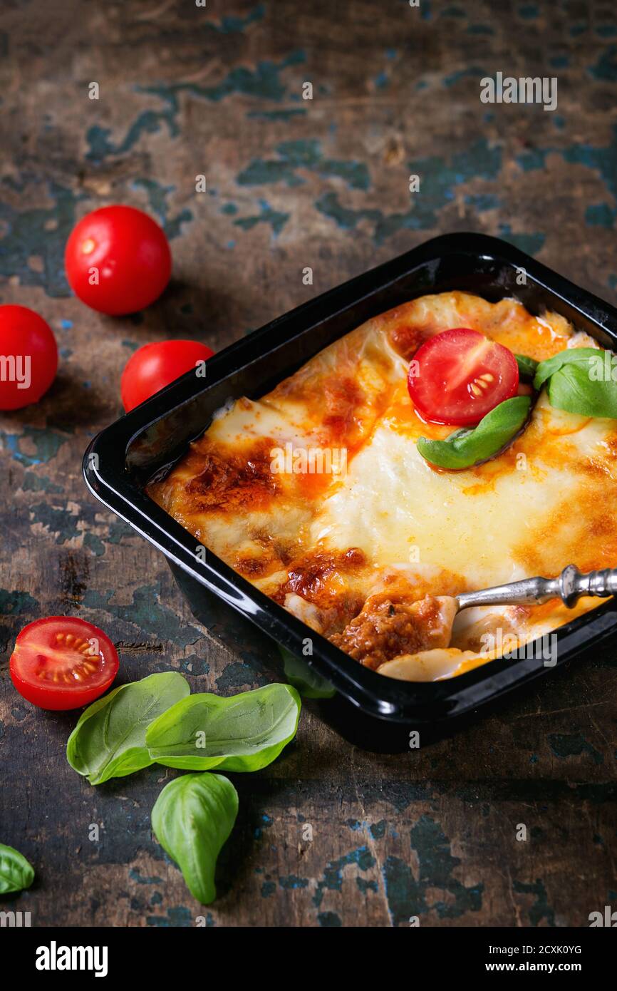 Lasagne di carne cotte in scatola di plastica nera, servite con forchetta vintage, pomodori ciliegini freschi e foglie di basilico su vecchio fondo testurizzato di legno scuro. Foto Stock