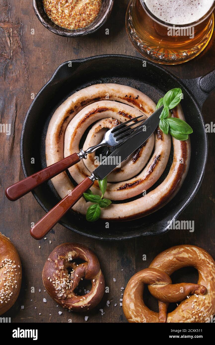 Bicchiere di birra chiara con salsicce fritte in padella con posate e tradizionali pretzel salati e la tazza di mostarda di oltre il vecchio legno scuro dello sfondo. Top vi Foto Stock
