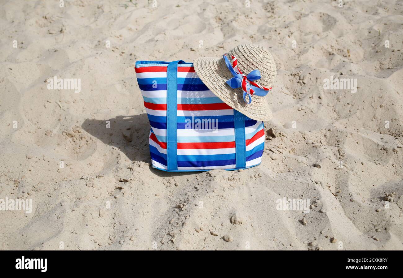 Colpo orizzontale con sabbia e cielo con accessori spiaggia femminile - borsa in tela a righe e cappello di paglia con nastri blu, bianco e rosso, francese, americano Foto Stock