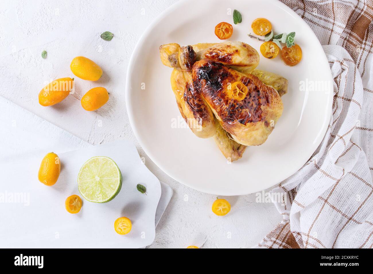Tutta la cottura di mini pollo arrosto con kumquat caramellati servita su piatto bianco con asciugamano da cucina, taglieri e agrumi freschi sopra chi Foto Stock